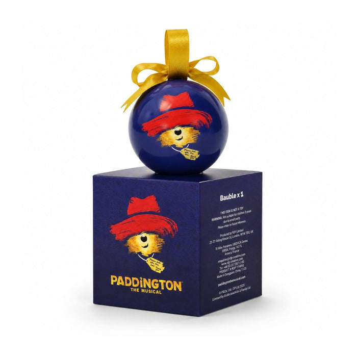 A blue bauble ornament with a gold ribbon loop and Paddington bear face wearing a red hat, displayed on top of a matching blue box labeled PADDINGTON THE MUSICAL.