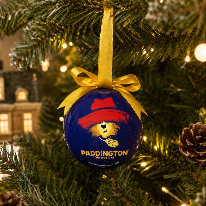 A round blue Christmas ornament hanging from a green Christmas tree with warm string lights. The ornament features Paddington the Bear wearing a red hat and has the text 'PADDINGTON THE MUSICAL' printed on it.