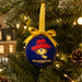 A round blue Christmas ornament hanging from a green Christmas tree with warm string lights. The ornament features Paddington the Bear wearing a red hat and has the text 'PADDINGTON THE MUSICAL' printed on it.
