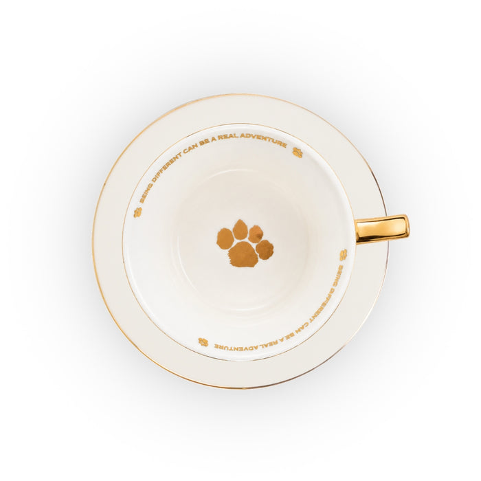 A white ceramic teacup and saucer set with gold trim, featuring a gold paw print in the center and gold text around the rim, shown from a top-down view on a white background.