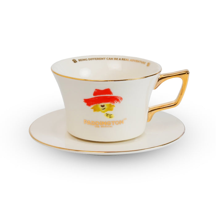A white porcelain teacup and saucer set with gold trim, featuring a red hat illustration and the text 'PADDINGTON THE MUSICAL' on the front, set against a plain white background.