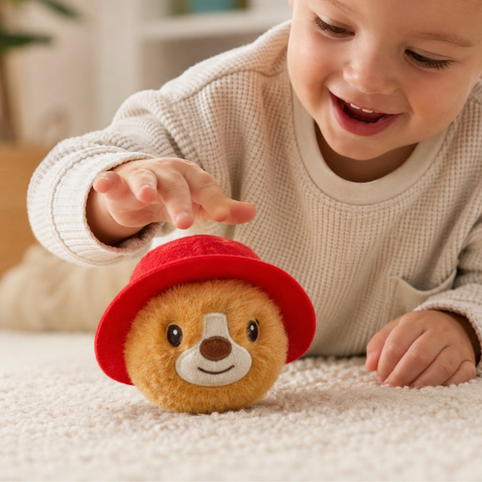 A toddler playing with a A round Paddington bear plush toy with golden-brown fur wearing a bright red hat with a yellow band, laying on a carpet
