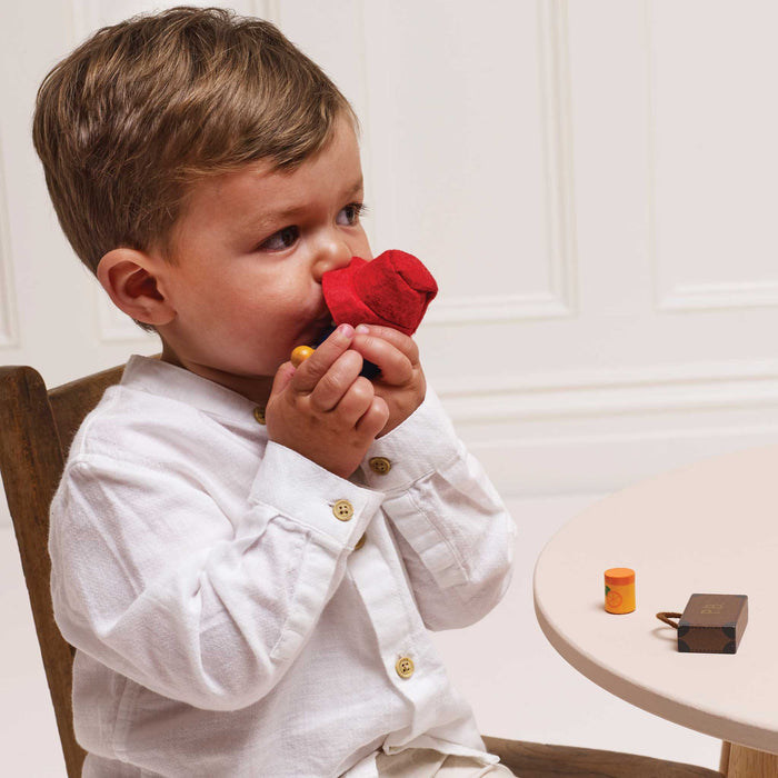 A toddler boy in a white long-sleeve shirt holds a red textured teething toy to his mouth while sitting on a wooden chair at a light-colored table.