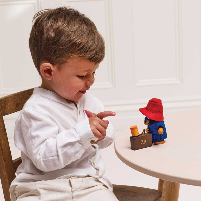 A toddler in a white long-sleeve shirt and beige trousers sits at a small round table, pointing toward a small Paddington-style toy figure with a red hat and brown suitcase.