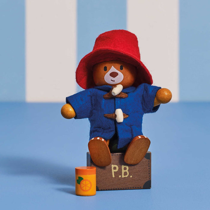 A toy Paddington Bear doll wearing a red hat and blue coat, seated on a small brown box labeled P.B.