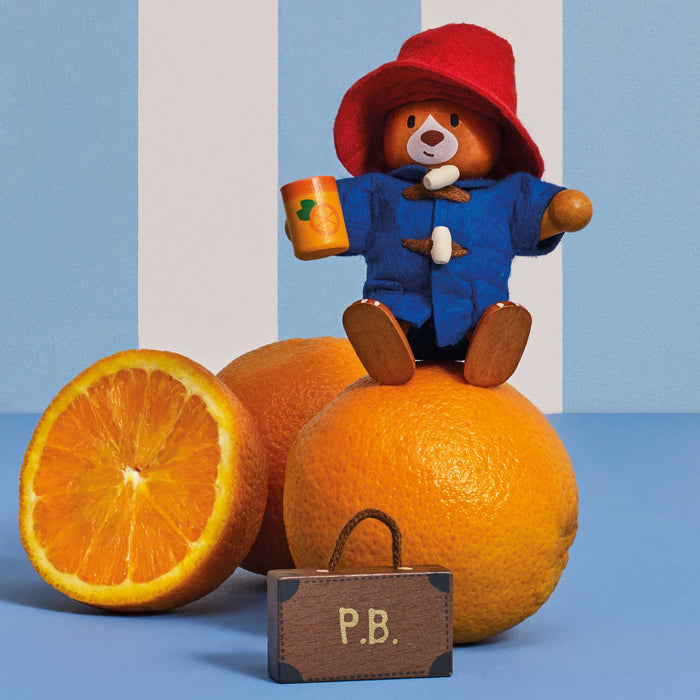A Paddington Bear wooden toy wearing its signature red hat and blue duffel coat, sitting on top of a whole orange and holding an orange mug. A cut orange half and a small brown suitcase labeled 'P.