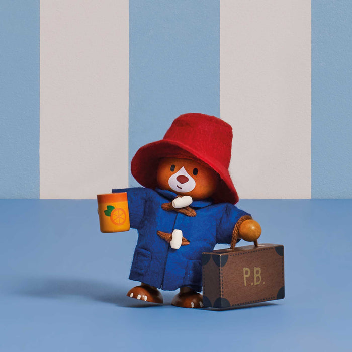A Paddington Bear collectible wooden figurine wearing a red hat and blue duffel coat, holding a yellow cup and a brown suitcase with 'P.B.