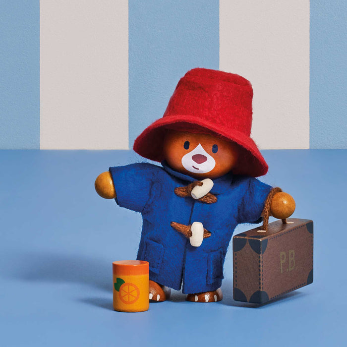 Paddington Bear dolls house figurine wearing a red felt hat and blue duffle coat, holding a brown suitcase and standing beside an orange tumbler, set against a blue and white striped background.