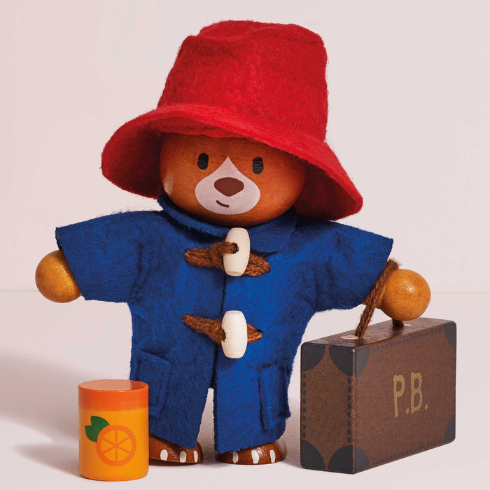 A wooden Paddington Bear dolls house toy wearing a red felt hat and blue felt duffle coat, standing beside a brown suitcase marked P.B.