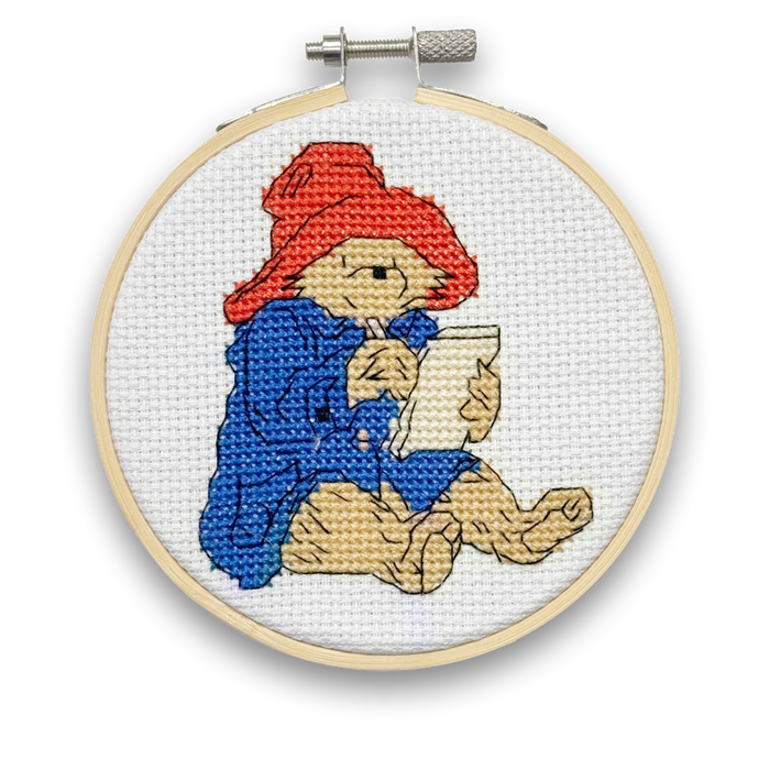 A mini cross stitch kit featuring Paddington Bear in a blue coat and red hat, sitting and writing a letter.