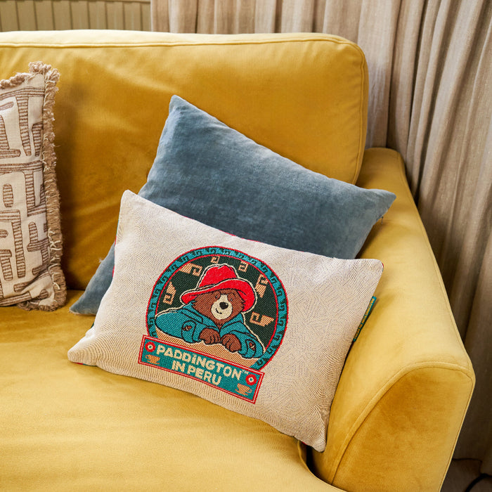 A cushion cover with an illustration of Paddington Bear wearing a hat, on a yellow sofa with other cushions.