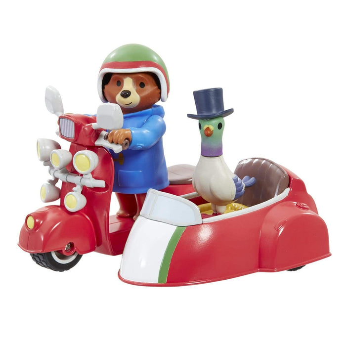 A toy set featuring a brown bear Paddington character wearing a green and red helmet and blue jacket, driving a red scooter with a sidecar. A white pigeon with a purple neck and black top hat sits in the sidecar.