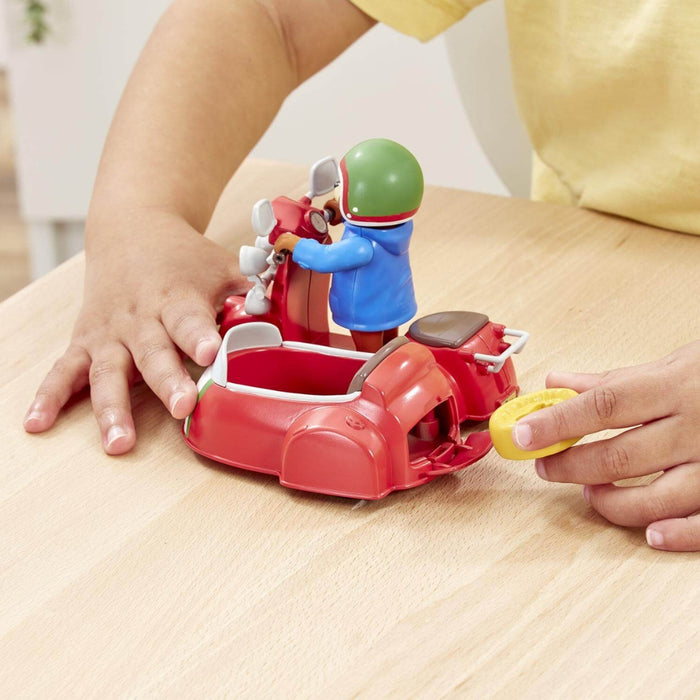 A child's hands play with a red Paddington toy scooter and sidecar set on a light wood table, with a person in a yellow shirt visible in the background.