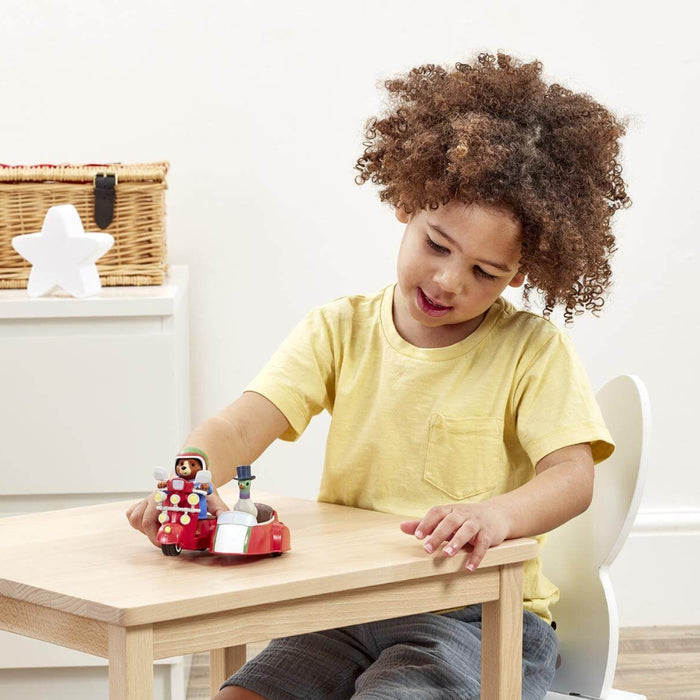 A young child with curly brown hair wearing a yellow t-shirt sits at a light wood table, playing with a small toy Paddington bear figurine set.