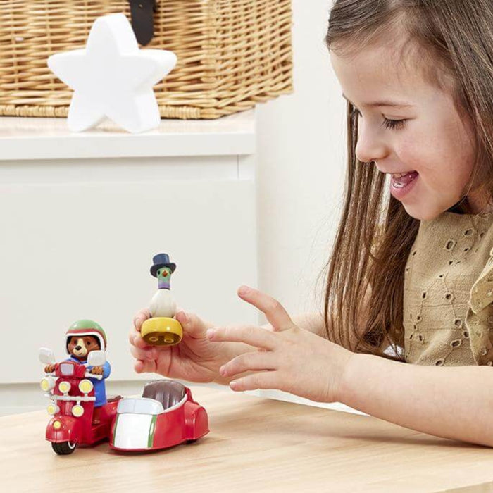 A young girl with long brown hair smiles while playing with a small Paddington bear toy set on a light wood table. The set includes a red sidecar with a Paddington figure and a small pigeon figure held in her hand.