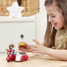 A young girl with long brown hair smiles while playing with a small Paddington bear toy set on a light wood table. The set includes a red sidecar with a Paddington figure and a small pigeon figure held in her hand.
