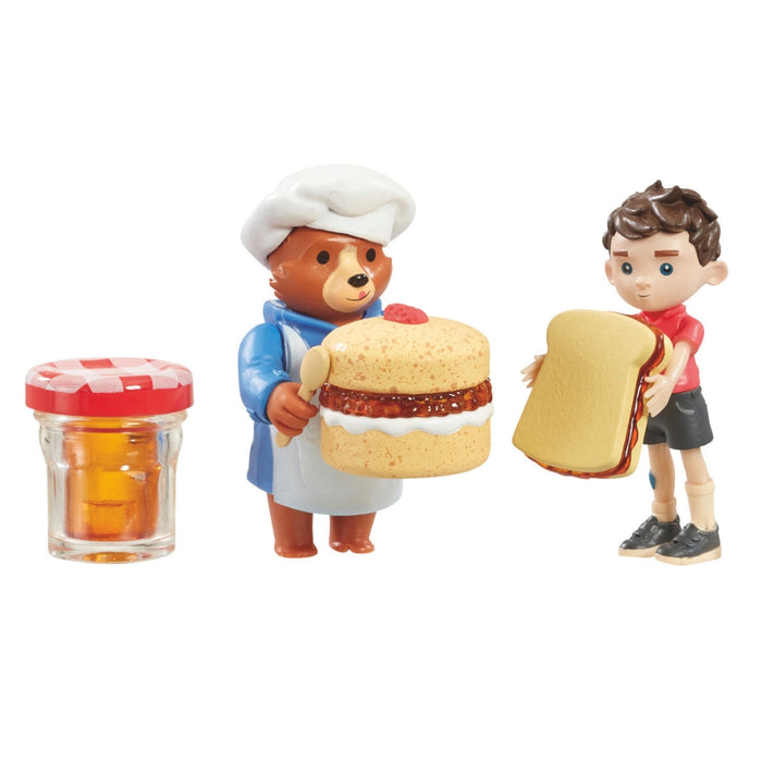Toy figure playset of Paddington bear in a chef's outfit holding a cake, a boy holding a sandwich, and a jar with a red lid on a white background.