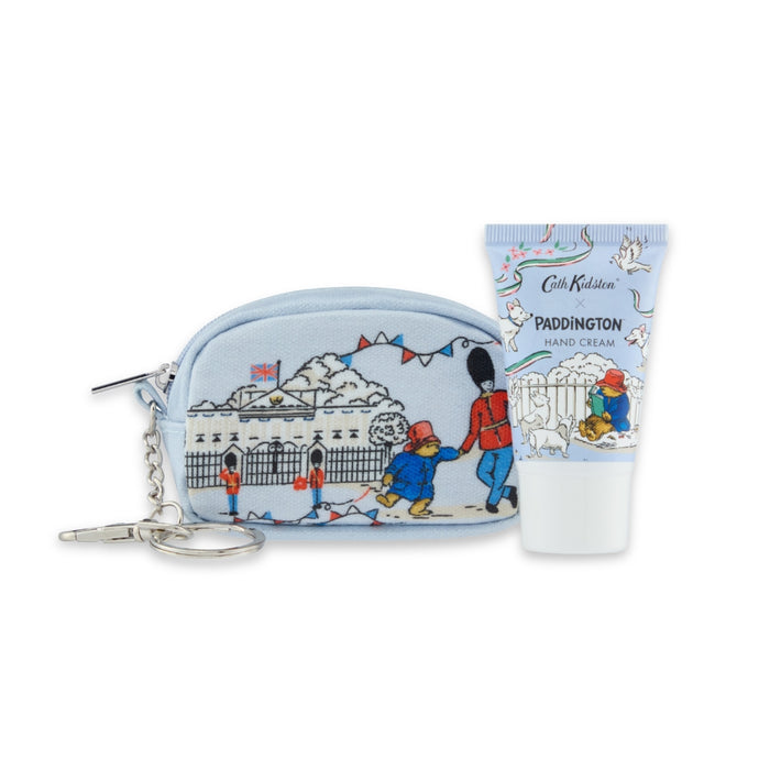 A light blue Paddington-themed zip pouch with a silver keychain, shown next to a matching light blue hand cream tube by Cath Kidston. The packaging features illustrations of Paddington Bear, a london soldier, and Buckingham Palace.