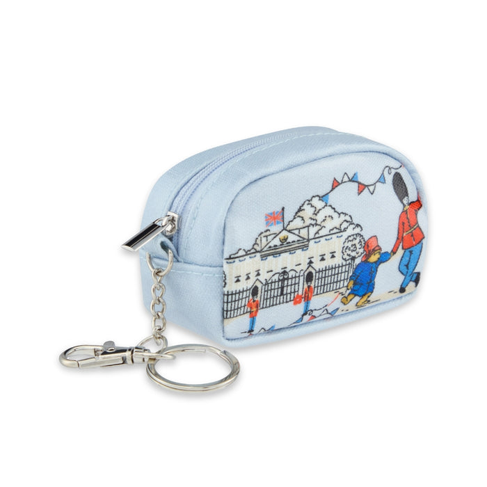 A light blue zippered coin pouch with a silver keychain clasp, featuring a printed illustration of Paddington Bear playing with a london soldier in front of Buckingham Palace.