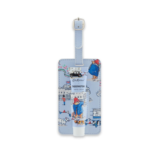 A light blue luggage tag featuring a colorful cartoon illustration of Paddington Bear and London-themed motifs, shown with a matching light blue tube of hand cream in front of it on a white background.
