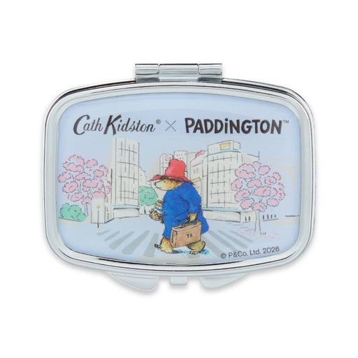 A rectangular compact mirror with rounded corners, featuring a silver metal frame and a light blue illustrated lid. The lid shows a cartoon scene of Paddington Bear walking with a suitcase, with the text 'Cath Kidston x PADDINGTON' at the top.