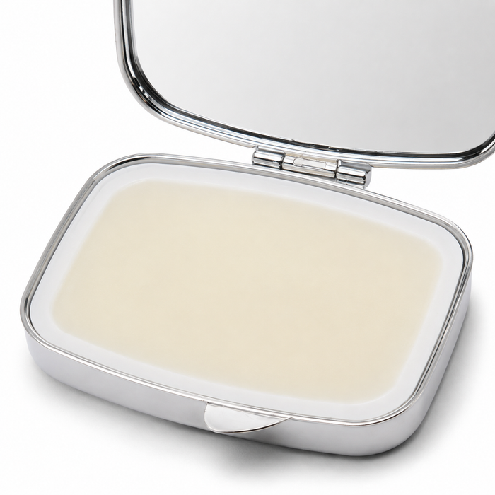 An open silver compact case containing a smooth, pale cream-colored balm or lip balm, with a mirror visible in the lid against a white background.