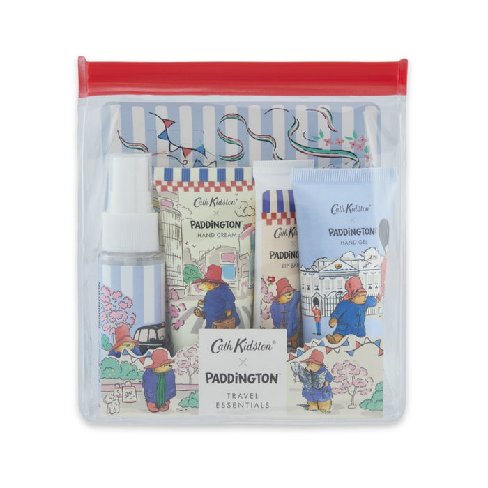 A clear zippered gift pouch with a red zipper seal, containing Cath Kidston x Paddington Travel Essentials items including a hand cream tube, lip balm, hand gel and boy mist, all with Paddington illustrations on a white background.