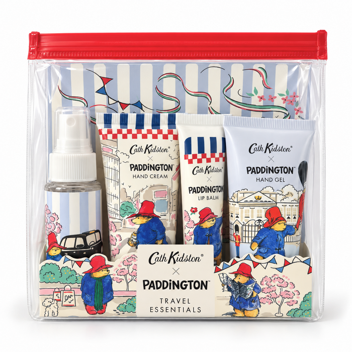 Cath Kidston x Paddington Travel Essentials gift set in a clear zip pouch with a red zipper. The set includes a hand gel bottle, lip balm, hand cream, and a hand gel tube, all decorated with Paddington illustrations and a central label reading 'Cath Kidston x PADDINGTON TRAVEL ESSENTIALS'.