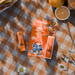 Paddington x Dr.PAWPAW Orange Marmalade Lip balm, featuring the main orange packaging card with Paddington illustrations, orange lip balm tube, and orange slices on a brown-and-white checkered picnic blanket.