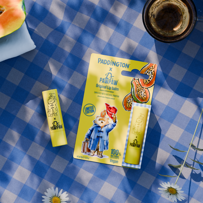 Paddington x Dr. Pawpaw Original Lip Balm displayed on a blue and white checkered picnic blanket, with a watermelon slice, a glass of iced coffee, and daisies as styled props.