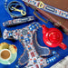 A flat-lay of Paddington-themed dog accessories on a bright blue background, including a harness, leash, and collar with blue-and-white gingham and red London bus motifs, alongside a brown suitcase, red teapot, cup of tea, and pastries.