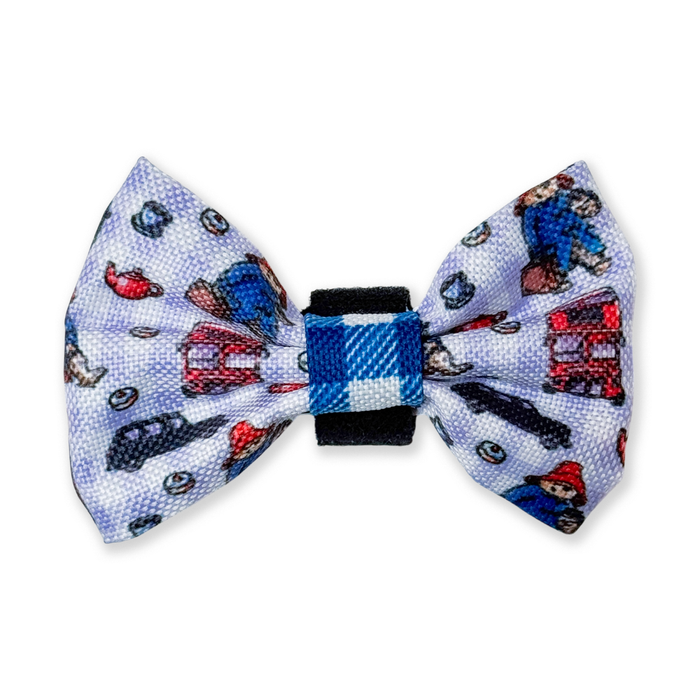 A small fabric bow tie featuring a light blue and white patterned design with paddington bear cartoon characters and london taxis, secured with a blue and white checkered center band and a black elastic closure.