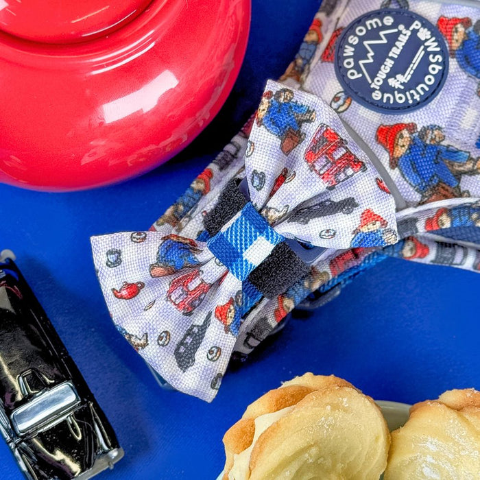 A fabric bow and matching headband featuring a colorful cartoon pattern of Paddington Bear, displayed on a bright blue surface alongside a red ceramic teapot, a toy car, and pastries.