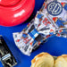 A fabric bow and matching headband featuring a colorful cartoon pattern of Paddington Bear, displayed on a bright blue surface alongside a red ceramic teapot, a toy car, and pastries.