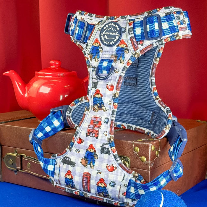 A dog harness featuring a blue and white gingham pattern with illustrations of Paddington Bear, red buses, and a red telephone booth, displayed on a brown suitcase with a red teapot and red background.