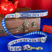 A Paddington-themed dog leash with a blue and white gingham strap, a printed pattern of red buses and red telephone booths, and gold-tone hardware, displayed in front of a brown vintage suitcase with red and blue props.