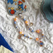 A colorful beaded phone strap with charms laid out on a white textured fabric surface next to a clear phone case featuring Paddington Bear illustrations.