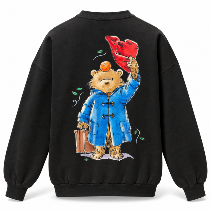 A black crewneck sweatshirt shown from the back, featuring a large graphic print of Paddington Bear in a blue duffel coat holding a red hat and a brown suitcase, with an orange on his head.