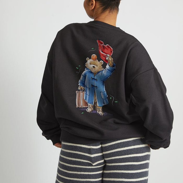 A child shown from the back wearing a black sweatshirt with a large graphic of Paddington bear in a blue coat holding a red hat and suitcase, paired with navy and white striped pants, against a light gray background.