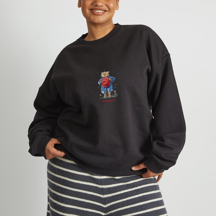 A person wearing a black crewneck sweatshirt with a small Paddington teddy bear graphic on the chest, paired with a navy and white striped knit top, against a plain light gray background.