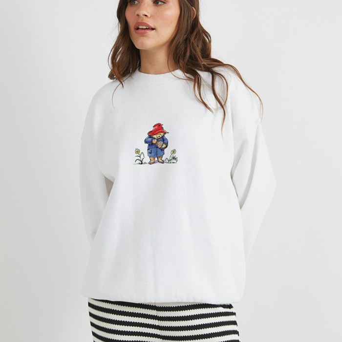A woman wearing a white crewneck Paddington sweatshirt with a small colorful cartoon character graphic on the chest, paired with a black and white striped skirt, against a plain light grey background.