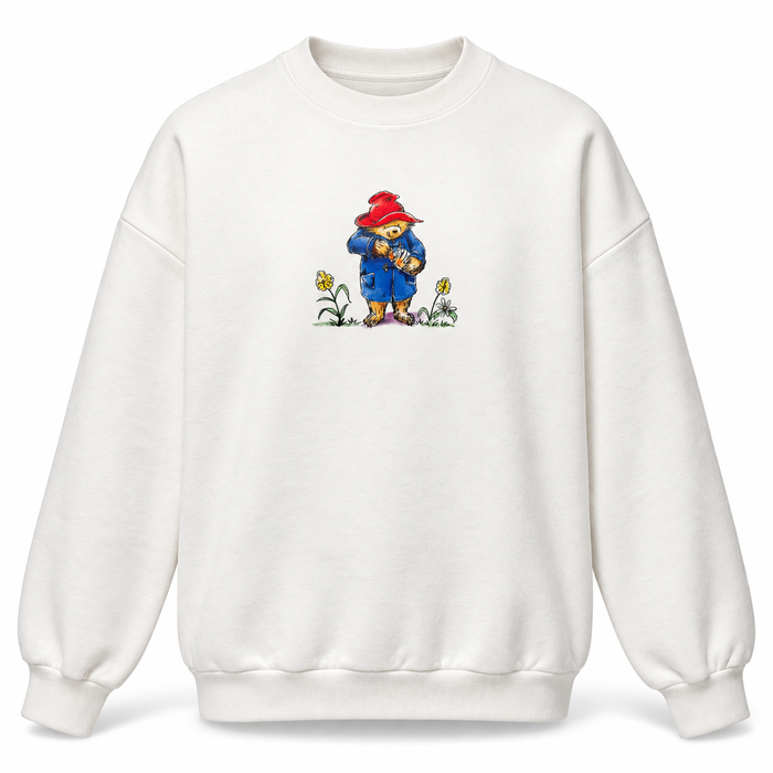 An off-white crewneck sweatshirt featuring a central graphic of Paddington Bear holding a jar of marmalade, with small yellow flowers at his feet, shown against a plain white background.