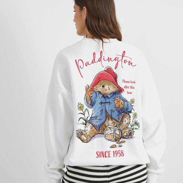 A woman shown from the back wearing a white sweatshirt with a large Paddington Bear graphic print. The design features Paddington in his signature red hat and blue duffle coat, with 'Paddington' written in red script above and 'SINCE 1958' below.