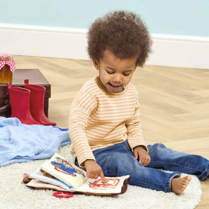 A toddler with curly hair sits on a white rug in a room, wearing a beige and white striped long-sleeve shirt and blue jeans, pointing at a soft play Paddington book.
