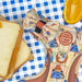 A dog harness laid flat on a blue-and-white checkered surface, styled with slices of toast and an orange slice. The harness has a multicolor cartoon print, a bow tie, and a circular patch reading 'Pawsome Paws Boutique Tough Trails'.
