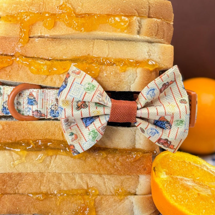 A close-up of a small fabric dog bow tie with a colorful illustrated pattern and orange-brown center, tied around a stack of sliced bread with orange jam, with whole and sliced oranges nearby on a warm brown background.
