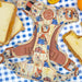 A dog harness featuring a colorful cartoon print of Paddington Bear on a cream background, styled on a blue and white checkered tablecloth with slices of bread and orange slices around it.
