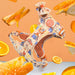 A dog harness with a colorful cartoon print, floating in a stylised studio composition with floating orange slices, toast, and marmalade against a peach background.