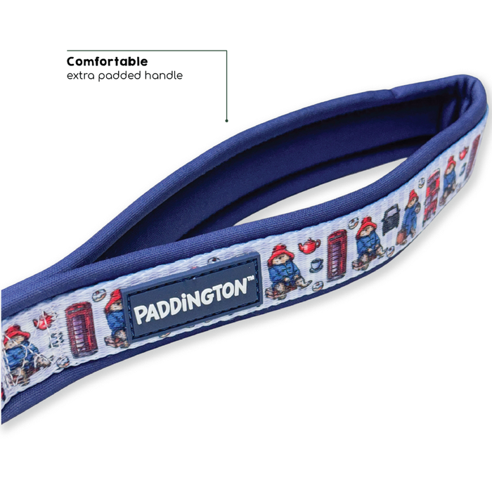 Paddington-themed padded handle strap for a dog leash, featuring a light blue patterned fabric with Paddington characters and red telephone booths, a navy blue padded handle, and a central black brand patch.