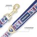 A close-up of a Paddington-themed dog leash with navy blue trim, a white patterned strap featuring red buses and blue bear characters, a gold-tone swivel clasp labeled 'locking clip twist base to lock,' and a gold D-ring. Text callouts explain the hardware, on a clean white background.